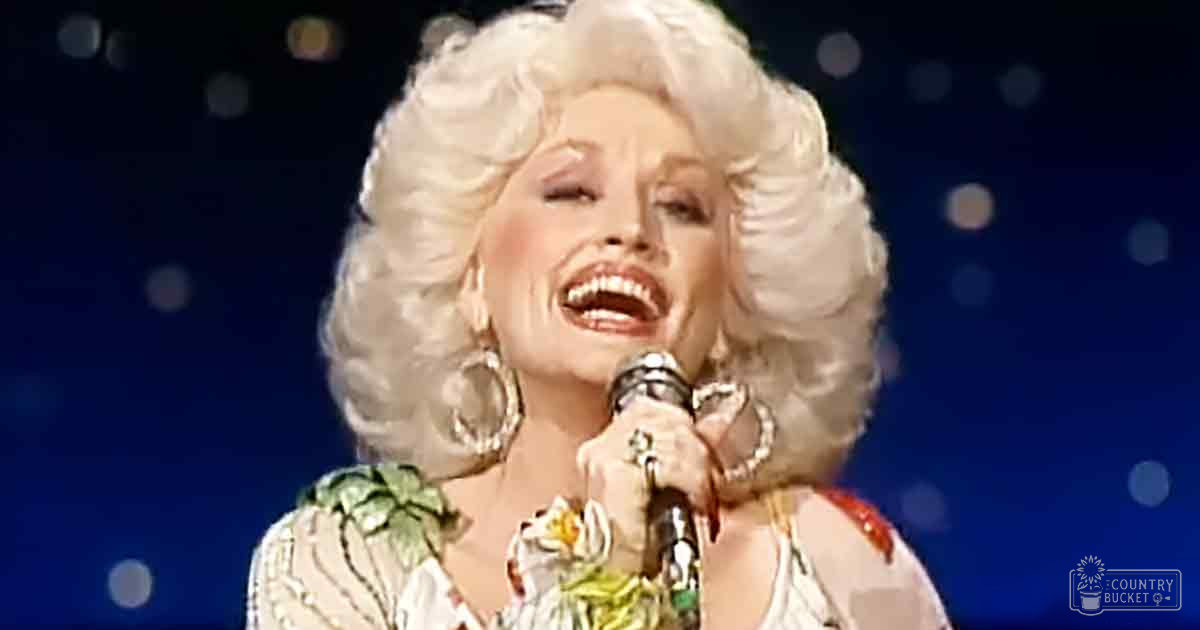 An adult female performer wearing a floral-patterned top in an indoor setting, holding a microphone with large hoop earrings and rings.