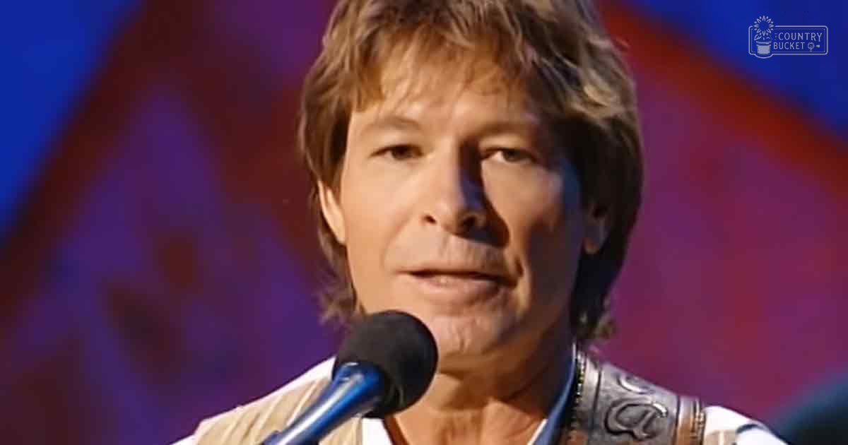 A middle-aged adult male wearing a white shirt with decorative patterns in an indoor performance setting with blurred stage colors.