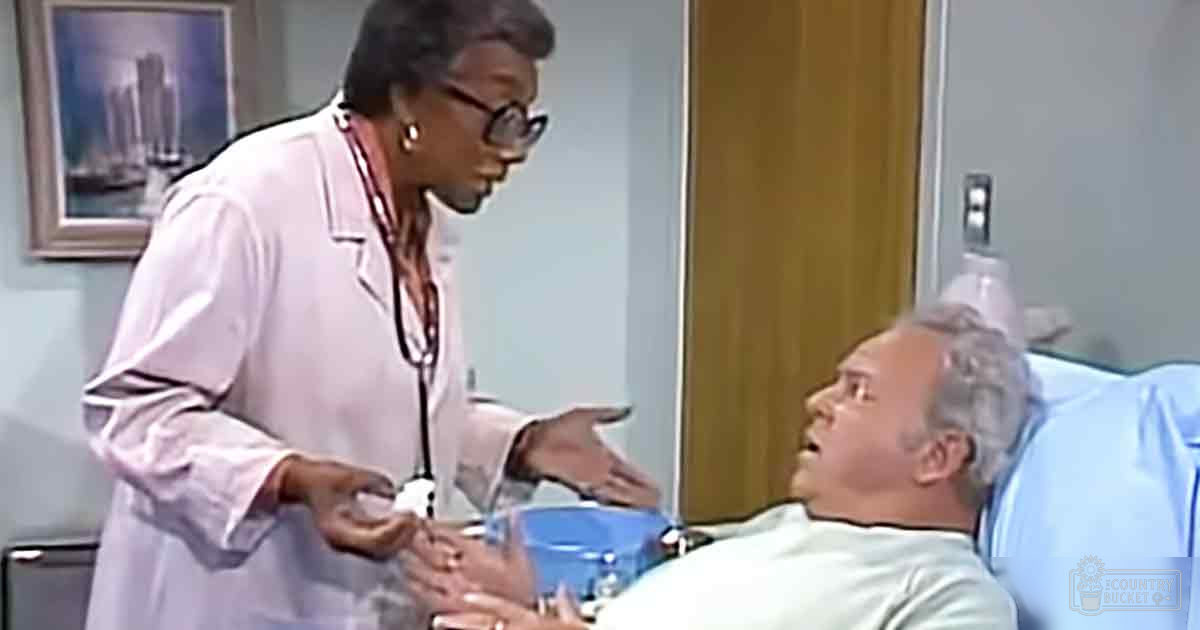 A middle-aged to older adult female healthcare professional in a white lab coat interacting with an older adult male patient wearing a light blue hospital gown in a hospital room, surrounded by medical furniture and home decor including a framed cityscape picture.