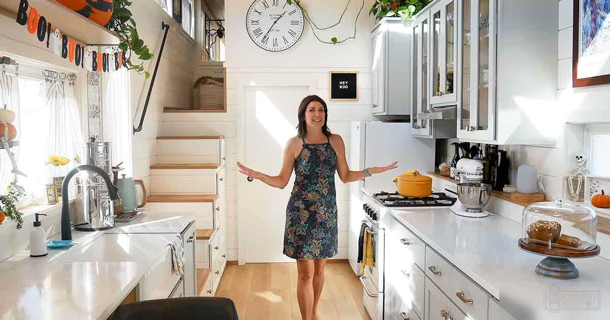 A young adult female in a floral dress with thin straps stands in a kitchen decorated for Halloween. The setting includes white countertops and cabinets, a gas stove, refrigerator, stand mixer, kettle, and various utensils.