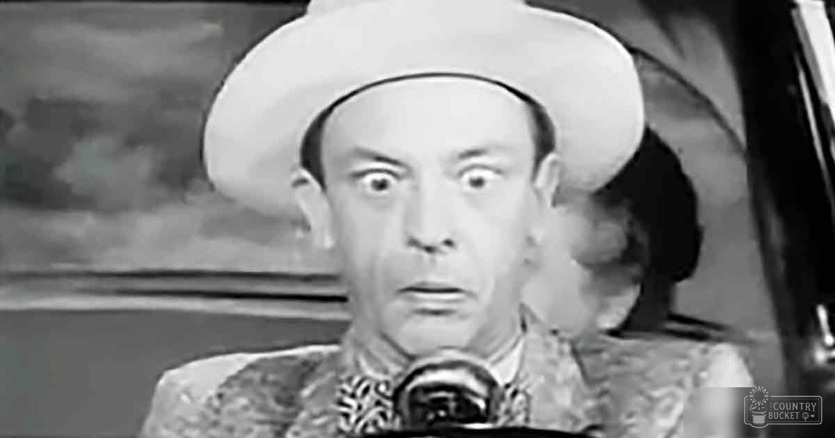 An older adult male wearing intricate traditional clothing with a wide-brimmed hat, set against an indoor stage or theatrical backdrop.