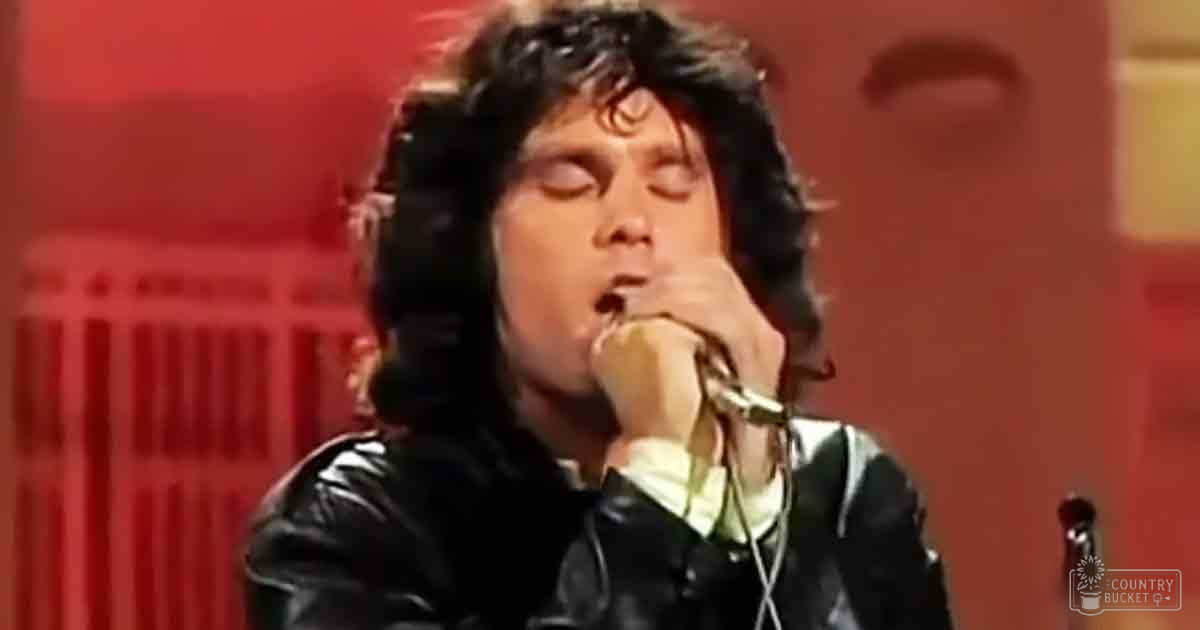 An adult male singer wearing a black leather jacket with high collar and full-length sleeves, holding a microphone in front of a red backdrop under professional lighting.