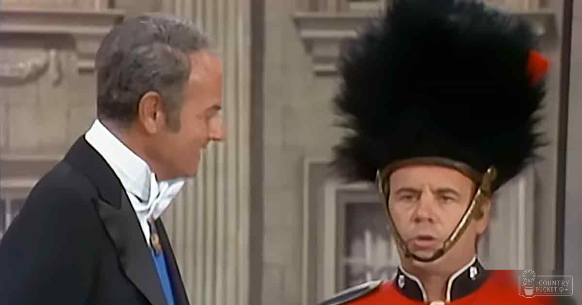 A middle-aged man in a dark suit jacket, white shirt with a bow tie, interacting with an adult male dressed as a historical British military guard wearing red uniform and tall bearskin hat indoors. The setting appears to be a well-lit, ornate official building from the late 19th century.