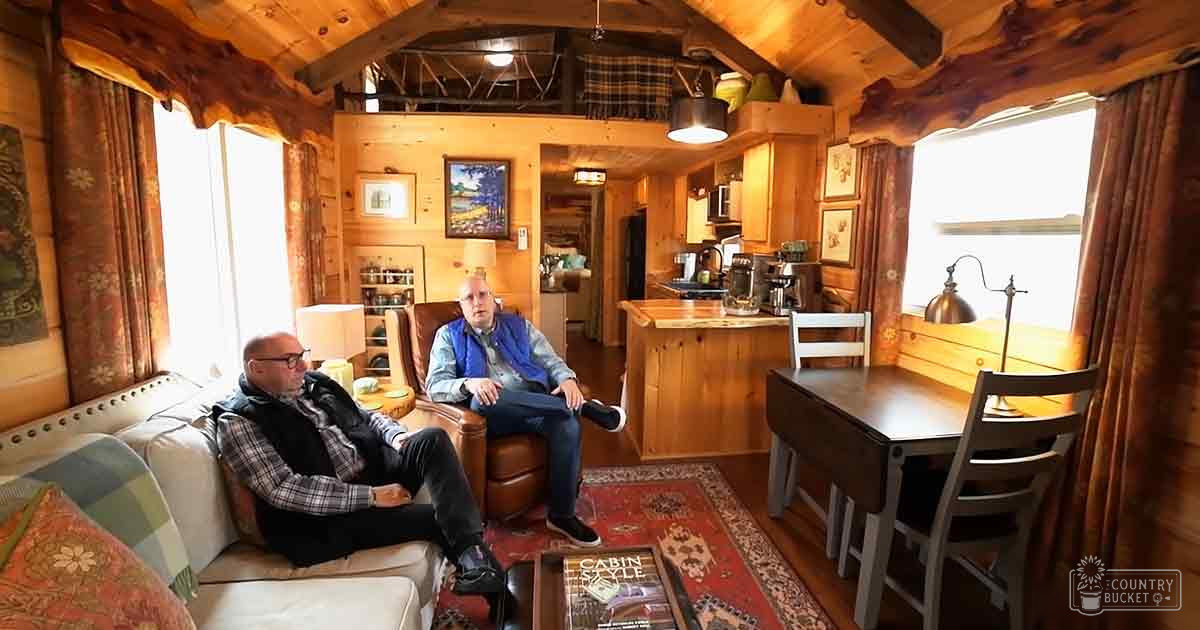 Two adults seated casually in a cozy cabin interior with wooden walls, large windows allowing natural light, and various cabin decor items like paintings, curtains, and lamps. The setting appears to be spring or summer based on the attire of the individuals.