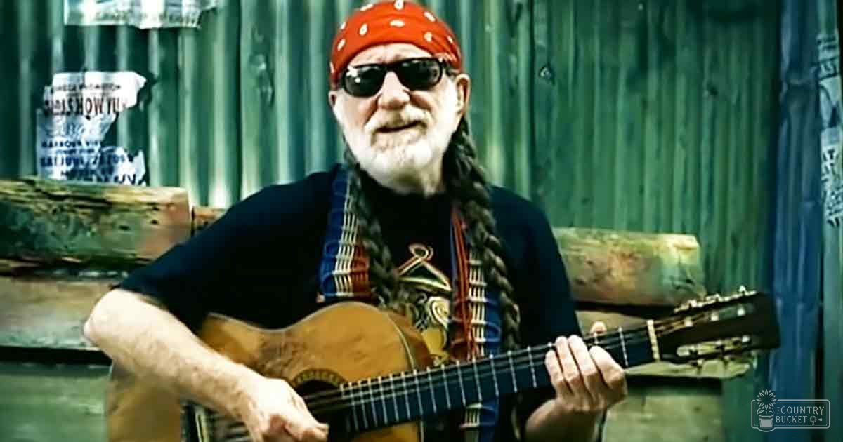 An older adult male playing an acoustic guitar outdoors near a wooden log bench with a can labeled 'SALSA SHOW' on it, under overcast or early evening light.