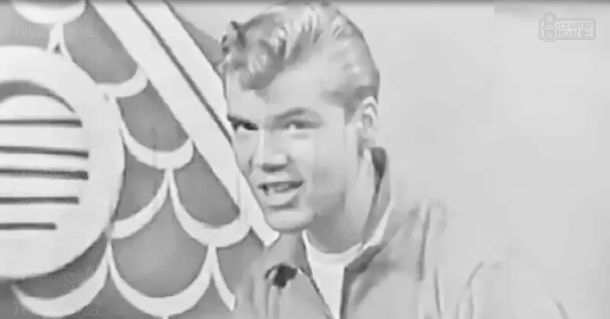 A young adult male wearing a light-colored collared shirt stands indoors, possibly during a performance or interview.