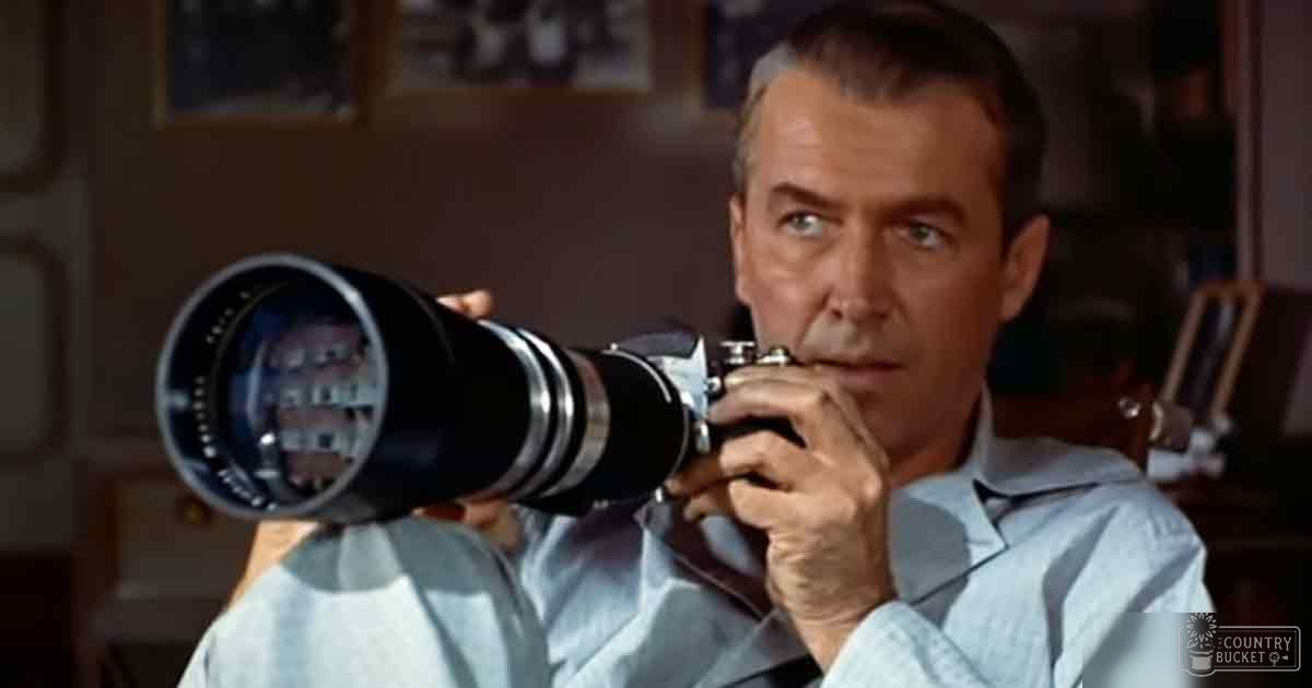 A middle-aged male wearing a light blue button-up shirt from mid-20th century, holding a large camera with a long lens in an indoor office or study setting.