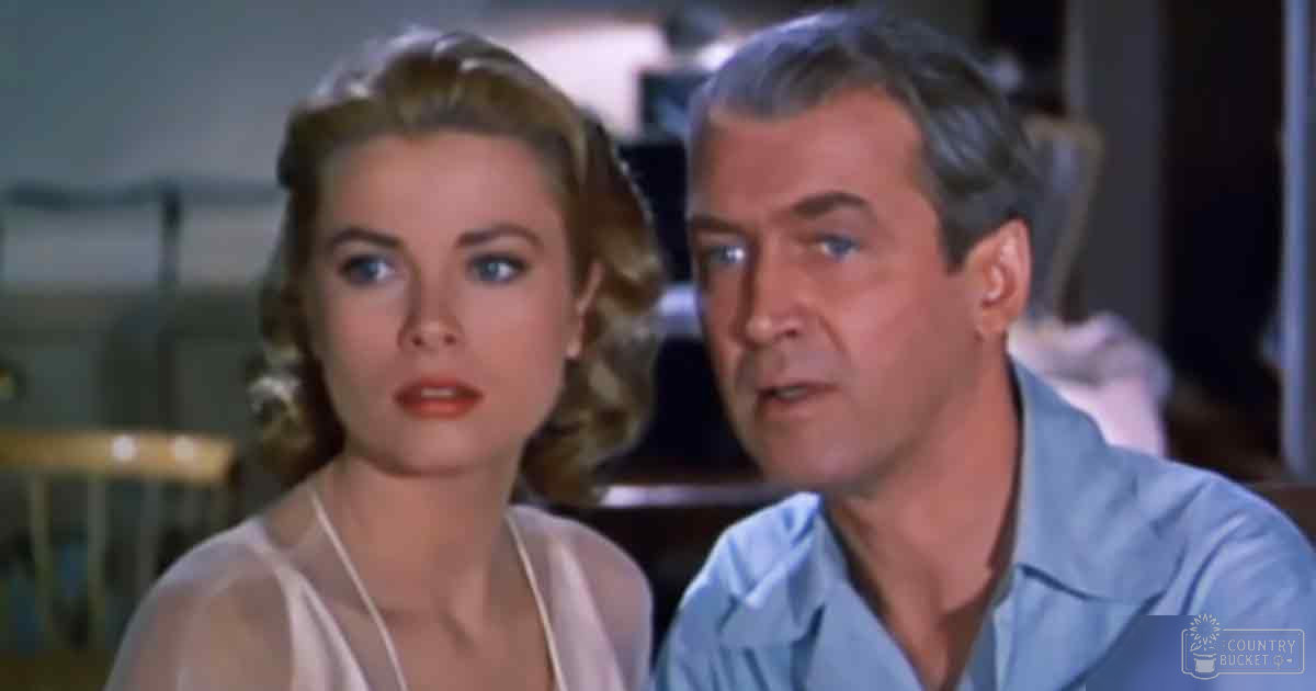 A mid-20th-century film set indoors features two individuals: a young adult female in a light-colored sleeveless top and an older male in a light blue collared shirt, surrounded by blurred furniture and decor elements.