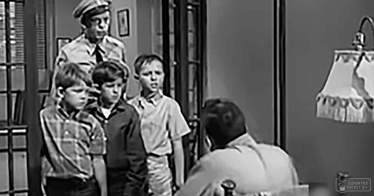 A group of children, likely male, wearing casual clothing indoors with a lamp and framed picture on the wall.