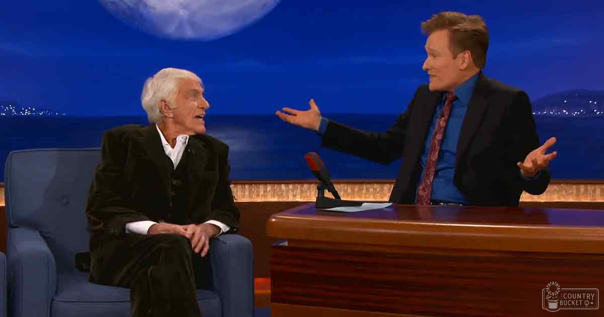 A talk show host in a dark suit jacket and patterned tie is interviewing an older adult guest wearing a black velvet blazer over a white collared shirt, set against a backdrop of a night sky with a full moon and city lights.
