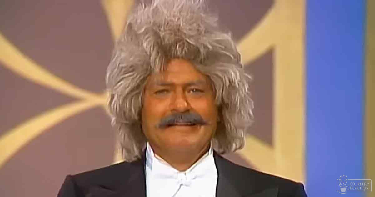 An older adult male in a black tuxedo with a white dress shirt and bow tie, set against an abstract geometric patterned backdrop suggesting a formal event.