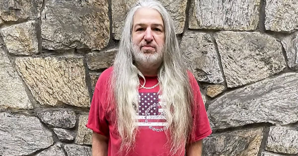 Middle-aged male in a red T-shirt with an American flag design, standing outdoors near a stone wall on a sunny day.