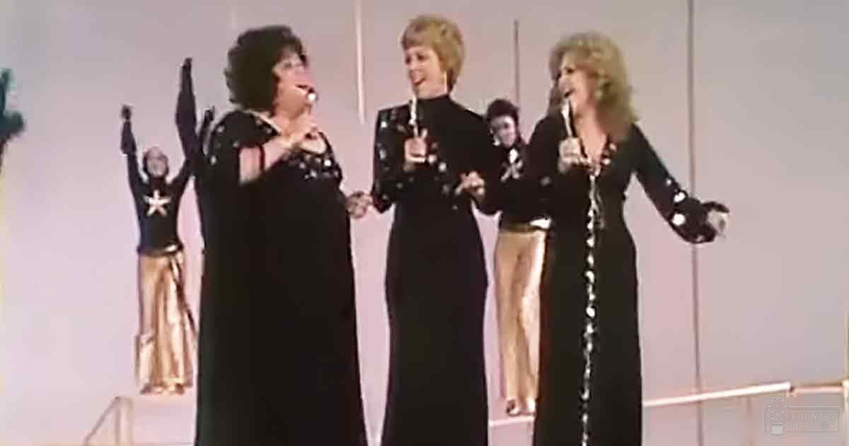A group of adult females in black dresses performing on a plain white stage backdrop under bright lighting, possibly during an indoor formal event.