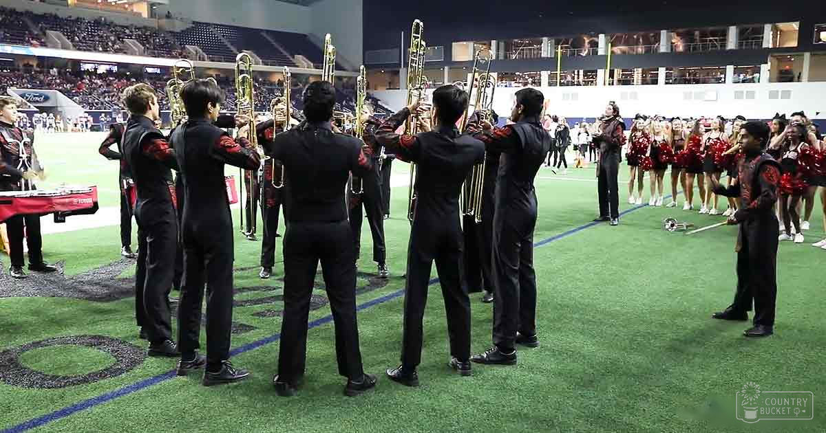 A marching band performing at an indoor stadium with young adults in black uniforms, red accents, and brass instruments; cheerleaders in sparkly red and black uniforms are on the sidelines.