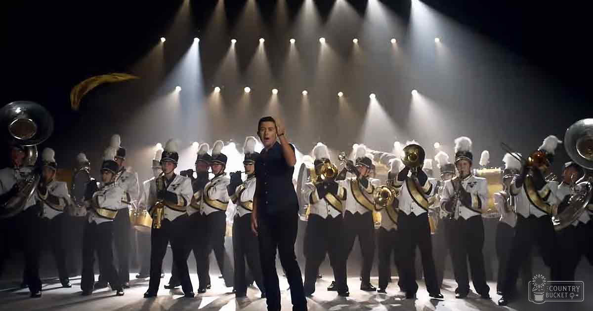 A marching band performance with a conductor leading young adult males in dark suits and white uniforms under bright stage lights in an indoor theater setting.