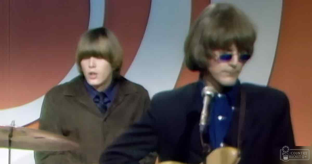 A musical performance scene with two male performers, one wearing a dark brown jacket over a blue shirt and the other in a dark suit jacket and blue shirt with small white dots, set against a colorful backdrop with abstract shapes. The setting suggests an indoor stage or performance area.