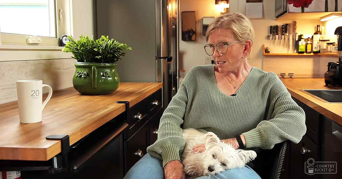 An older adult female wearing a green ribbed sweater in a kitchen setting with a small white dog nearby.