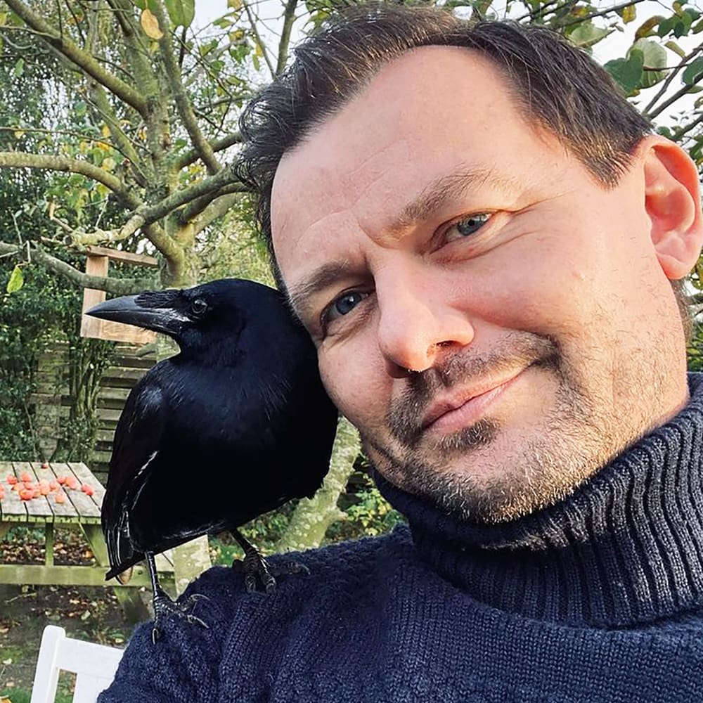 A middle-aged adult male stands outdoors with a black crow perched on his shoulder, surrounded by an autumnal garden scene with scattered leaves and trees.