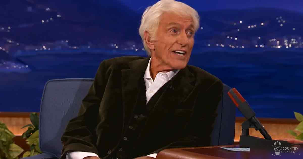 An older adult male in a studio setting with dark blue chair and nighttime cityscape background, wearing a dark blazer over a white shirt.