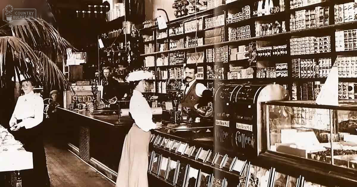 Victorian-era general store interior with adults in formal attire, likely from the late 19th or early 20th century; counter and shelves filled with various goods, including seed and coffee signs.