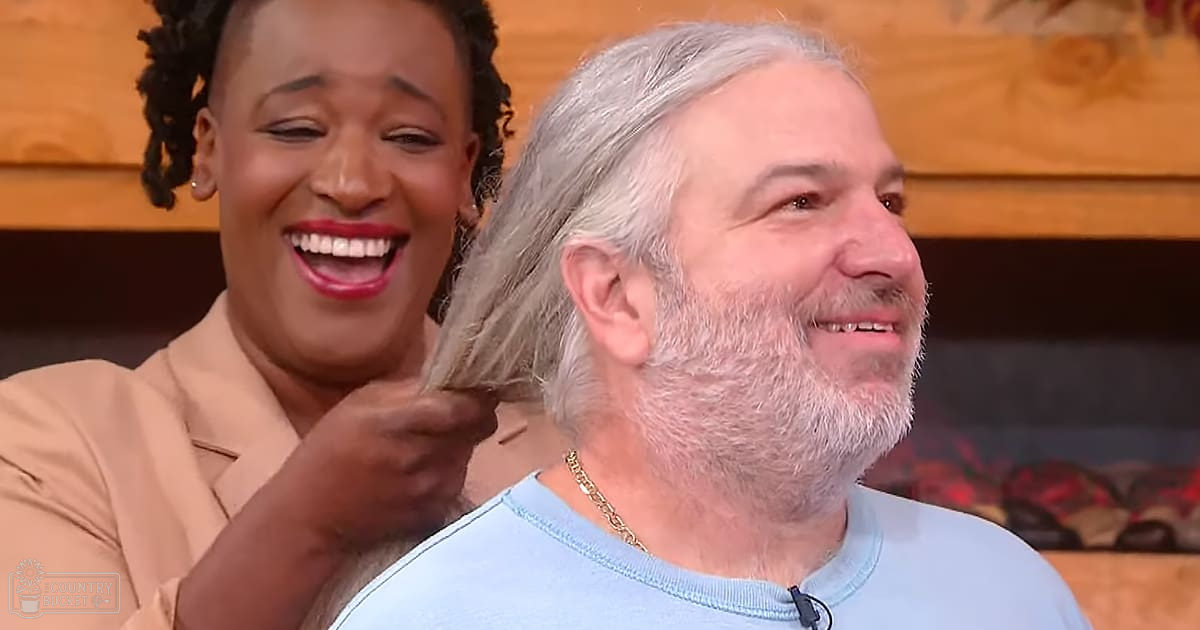 An adult male with gray hair and beard, wearing a light blue crew-neck t-shirt, interacts in an indoor setting with another individual dressed in a beige blazer or jacket with braids or twists. The background suggests wooden paneling and possibly a fireplace area.