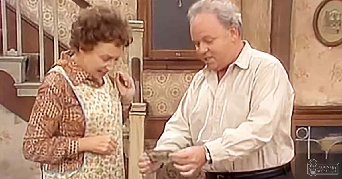 An older adult woman in a patterned dress and an adult male wearing a long-sleeved shirt, seated indoors in what appears to be a home setting with vintage furniture and decor.