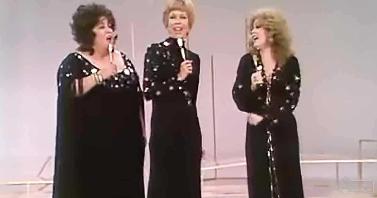 Three women singing in a studio setting, dressed in 1970s-style black dresses adorned with white dots or stars and silver sequins.