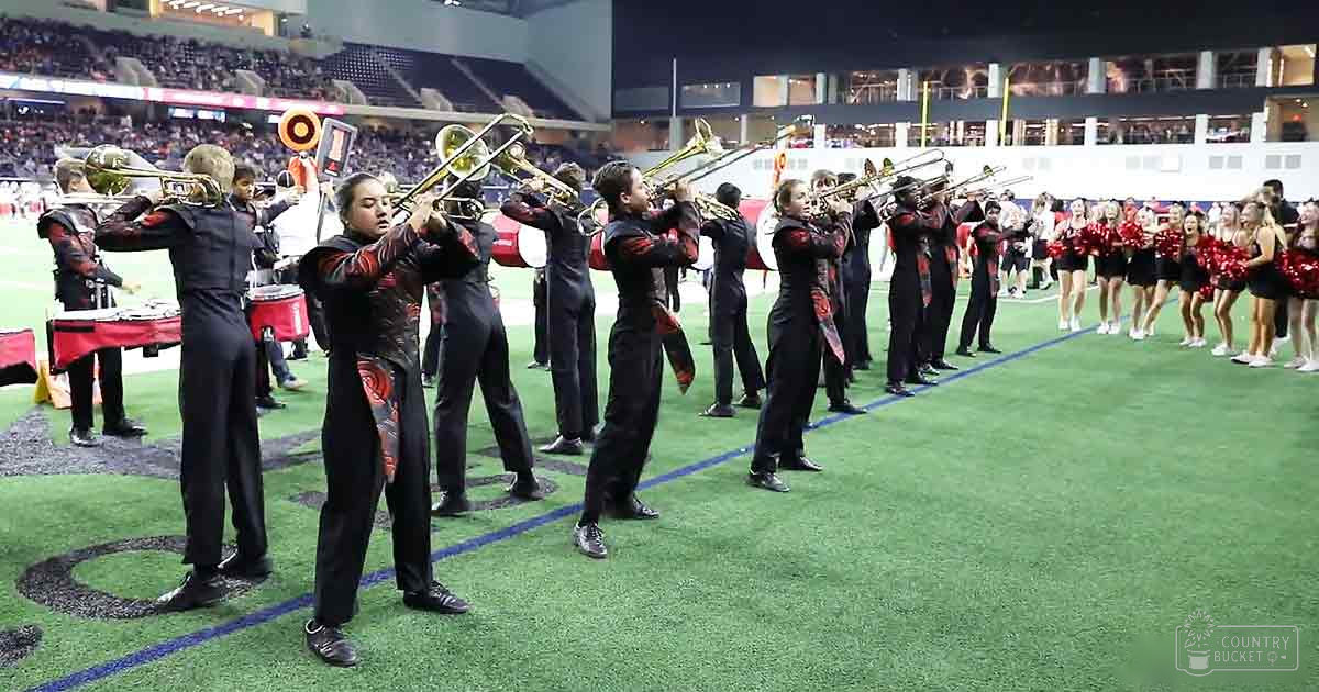 A marching band at a high school halftime show in an indoor stadium with cheerleaders holding pom-poms, surrounded by spectators.