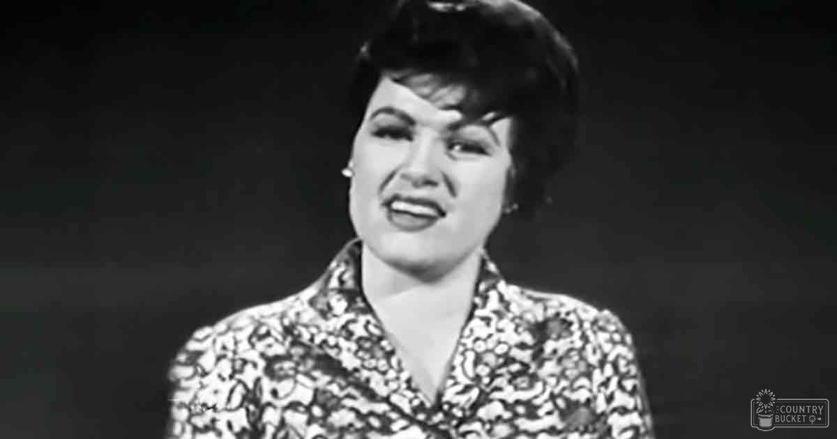 Middle-aged adult female wearing a floral blouse with short sleeves, V-neck style, and small earrings against an indoor backdrop suggestive of a performance or interview setting.
