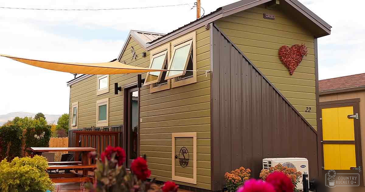 A tiny house with a yellow awning, small windows with open shutters, heart-shaped wreath, wooden picnic table, potted plants, and an air conditioning unit in an outdoor setting with greenery and overcast daylight.