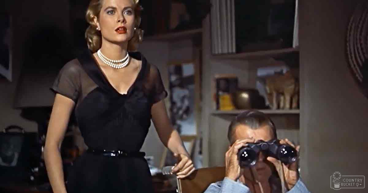 An adult female wears a fitted black dress with short sleeves and a low-cut neckline, accessorized with a pearl necklace; an adult male holds binoculars in the background of a room filled with shelves and furniture.