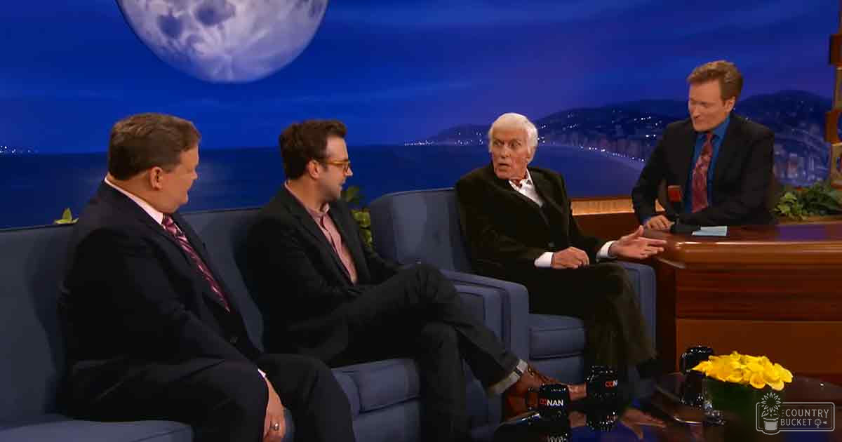 Talk show set in a studio, evening backdrop with hills and lights, blue armchairs, moon-like image in sky, professional setting with no personal items visible.