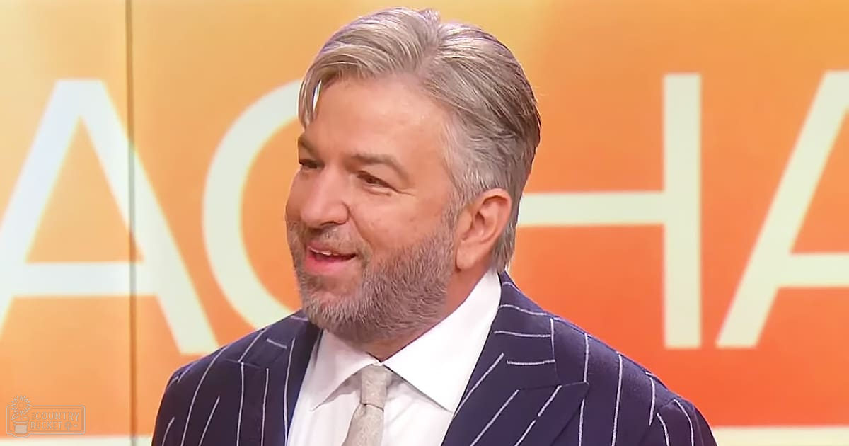 Middle-aged man in a dark blue pinstriped suit jacket and collared white shirt on an orange and white backdrop in a television studio setting.