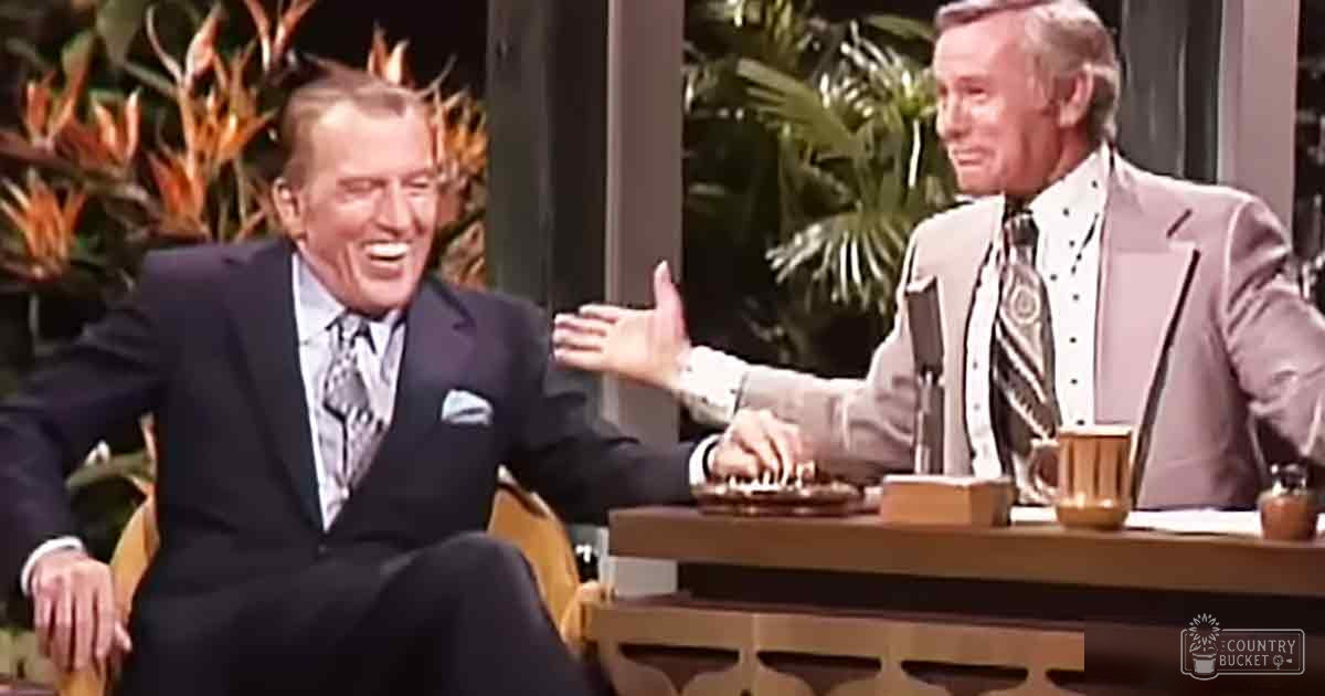 Two middle-aged men in formal suits with patterned ties, seated at a wooden desk in an indoor studio setting from the 1970s or early 1980s, surrounded by decorative plants.