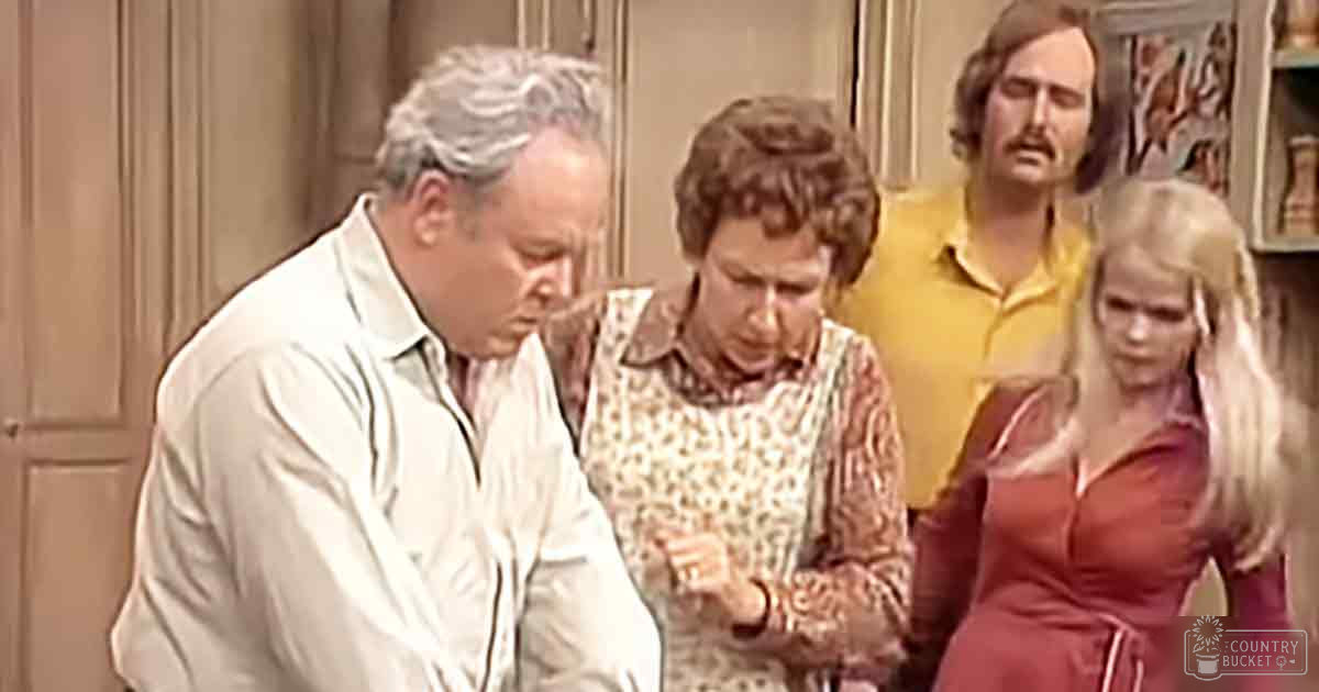 Two women in vintage floral dresses, a man wearing a light jacket over a button-up shirt with a patterned design, and another man in a yellow shirt, set against wooden furniture in an indoor room. Text 'Madly Odd' on the red dress.