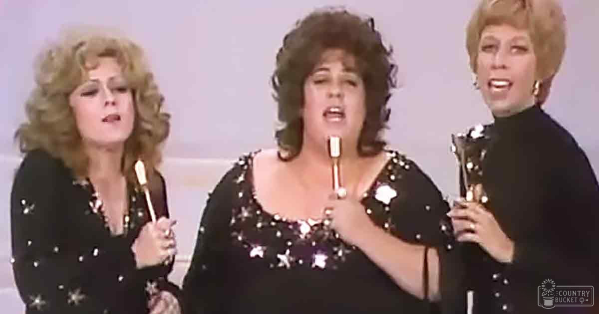 An adult female singer wearing a black sequined dress with silver stars, accompanied by two other women in black outfits holding microphones and an award on stage during an awards ceremony.