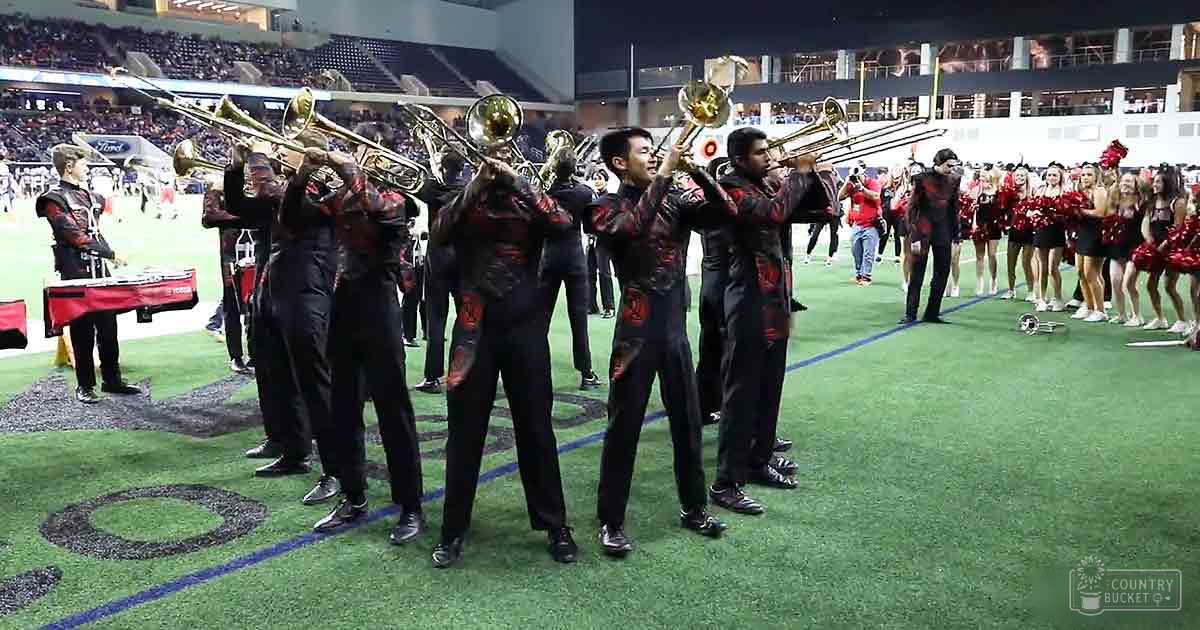 A marching band performs at an indoor stadium with football fields visible in the background; spectators sit on bleachers filled with artificial turf under daytime light.