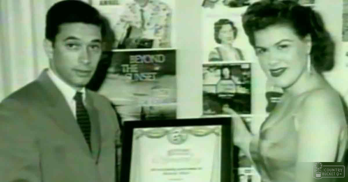 A young adult male in a light-colored suit jacket with a darker tie stands beside an adult female wearing a halter-style top, both indoors, with a framed certificate and 'Beyond the Sunset' book visible on a shelf behind them.