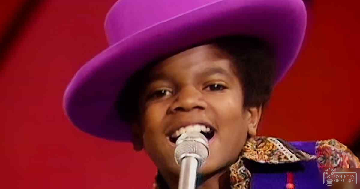A young adult male performer wearing a wide-brimmmed purple hat and a patterned shirt or jacket with floral designs stands on stage in front of a red background.