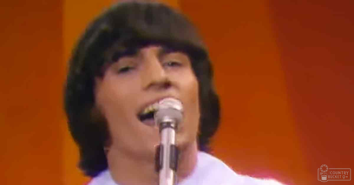 A young adult male singer wearing a white t-shirt and dark beanie, holding a microphone against an orange stage backdrop.