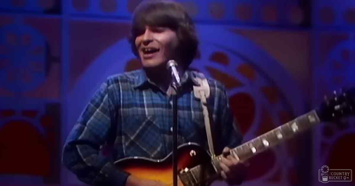 A young adult male singer performing on a stage in an indoor venue, wearing a plaid shirt from the late 1960s to early 1970s.