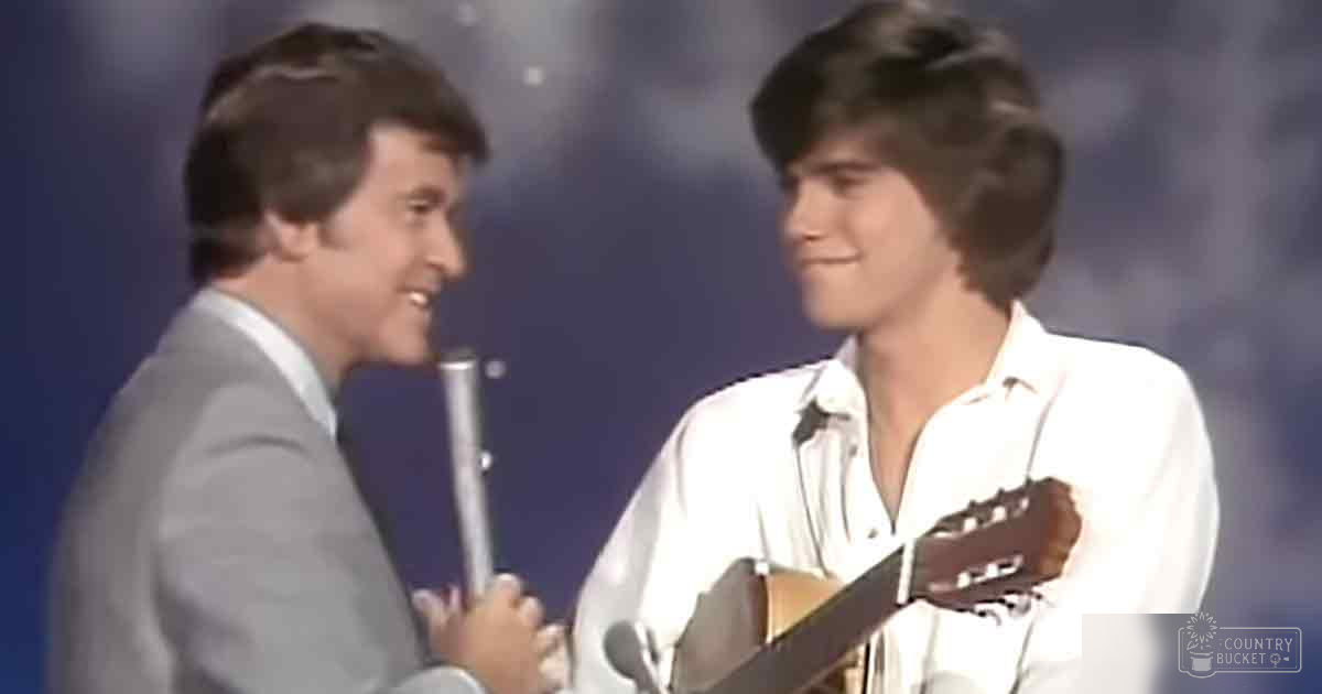 A young adult male plays a guitar on stage, accompanied by an older individual holding a microphone in the background.