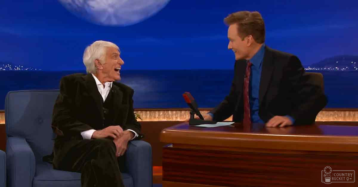 A male host and older adult guest are seated on stage during a late-night talk show interview; the host wears a dark suit with blue shirt and red tie, while the guest is in a black suit jacket over a white collared shirt. The setting includes a blue armchair for the guest and a wooden desk with a microphone, papers, and small object for the host against a backdrop of a large moon and night sky over water.