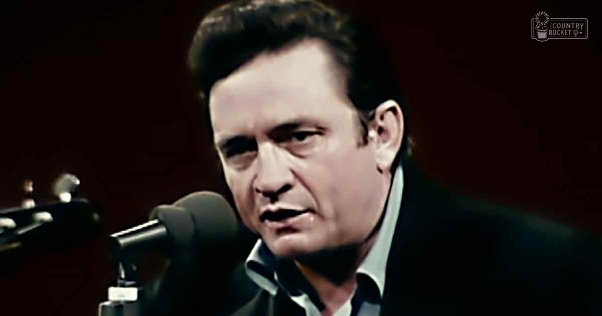 A middle-aged man wearing a dark suit jacket over a collared shirt with 'Madly Odd' text in the top right corner, performing on stage or in a studio setting.