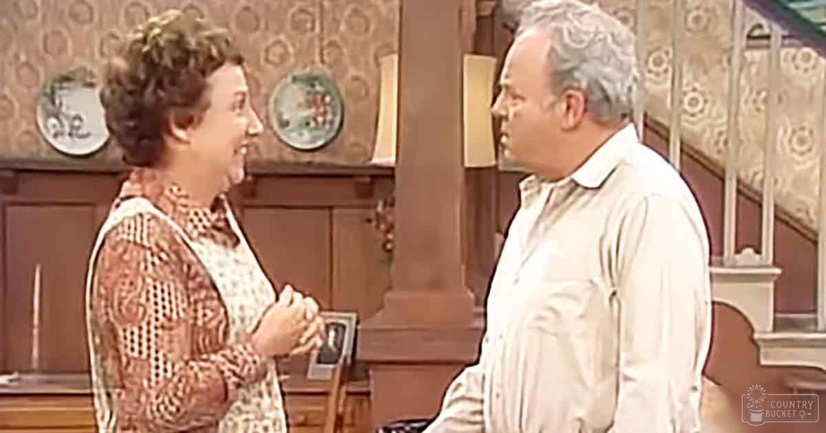 Middle-aged woman in patterned blouse and middle-aged man in striped button-up shirt, indoors with wooden furniture and decorative plates on mid-20th century wallpaper.
