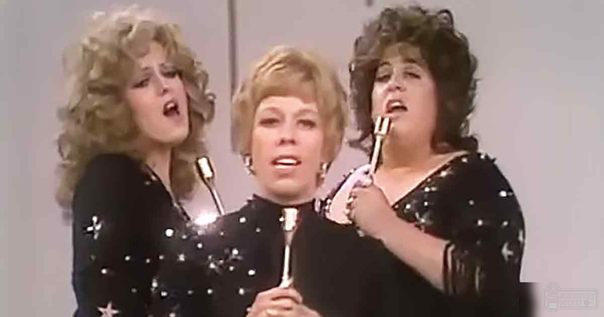 A musical performance scene with three adult females in matching black star-patterned outfits, holding microphones. The setting appears to be indoors with a plain background.