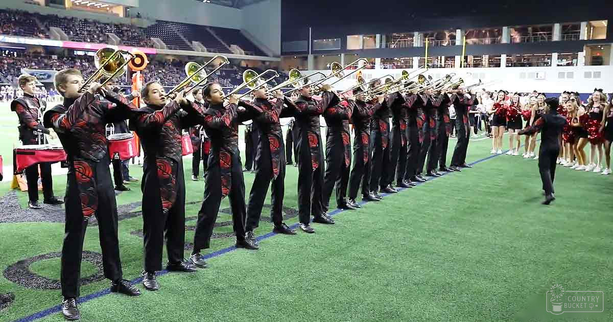 A marching band performing at an indoor stadium filled with spectators under artificial evening lighting.