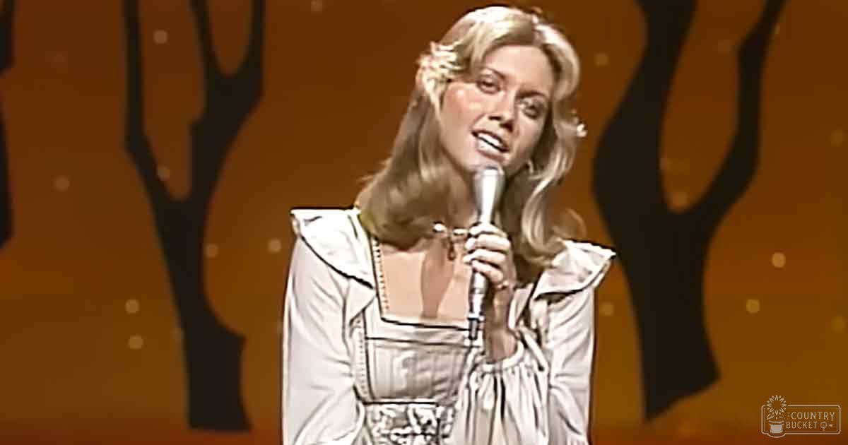 An adult female performer in a white, patterned dress with puffed shoulders and long sleeves on stage under artificial lighting.
