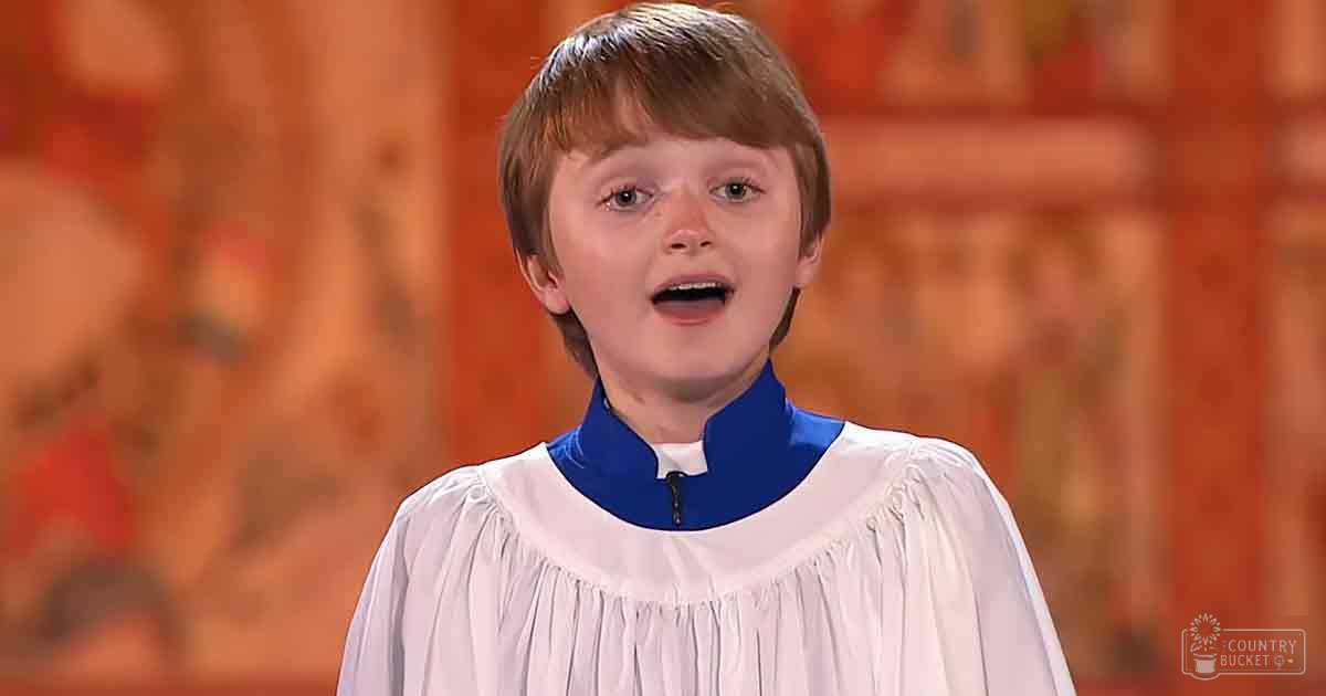A young child wearing a white and blue choir robe stands indoors within what appears to be a church setting with wooden paneling and potential religious artwork in the background, illuminated by natural daylight.