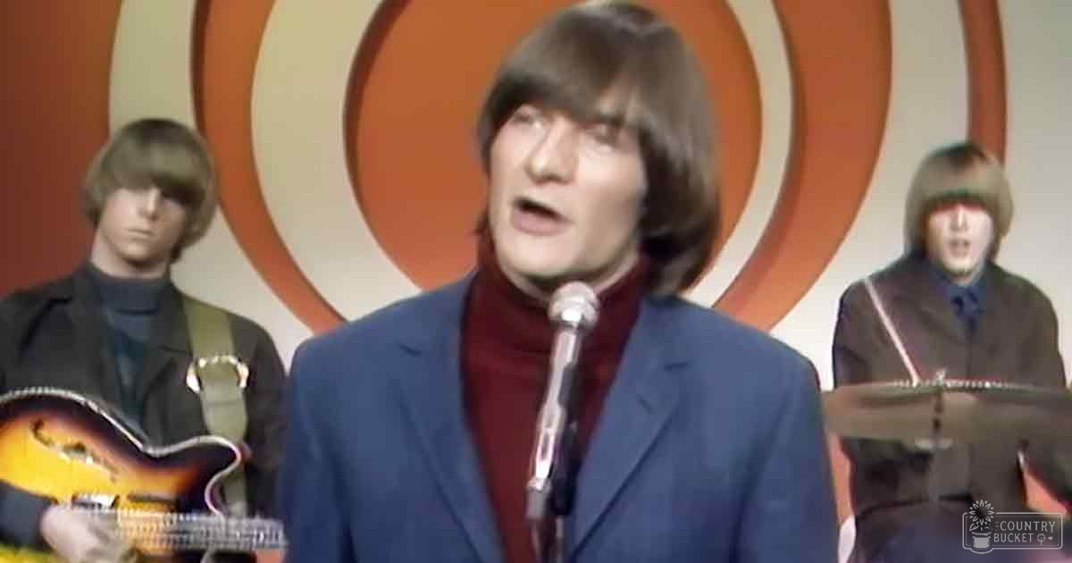 Young adult male in a blue blazer over a red turtleneck, singing into a microphone with background musicians in similar mid-20th century attire on a studio set designed for music videos.